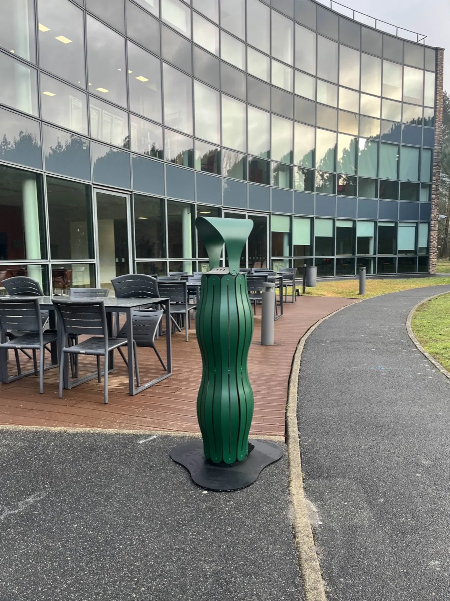 Cendrier vert sur pied au bord d&rsquo;une all&eacute;e de campus, pr&egrave;s d&rsquo;une terrasse