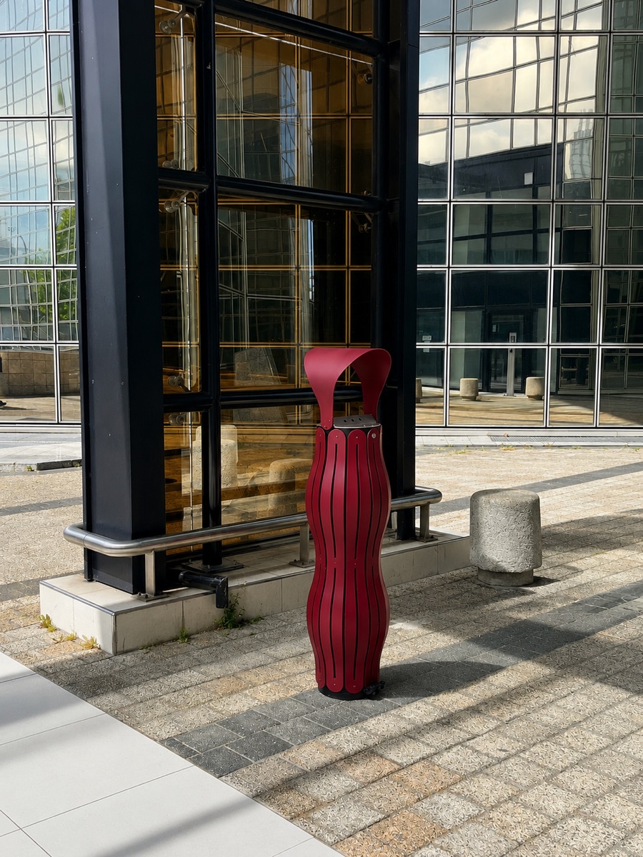 Cendrier rouge vigne pos&eacute; sur un parvis pav&eacute;, devant une fa&ccedil;ade vitr&eacute;e