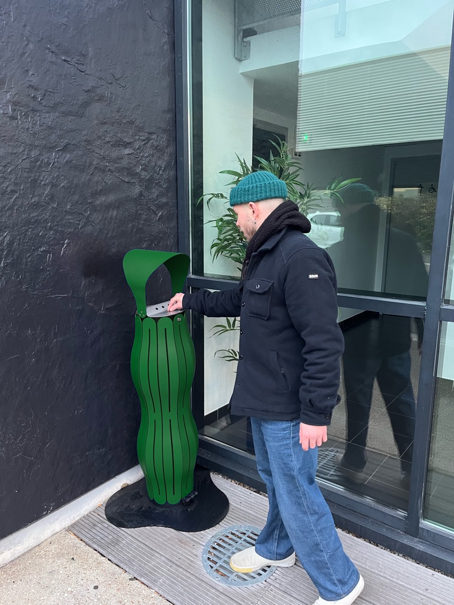 Un collaborateur utilise le cendrier vert devant un b&acirc;timent de bureaux